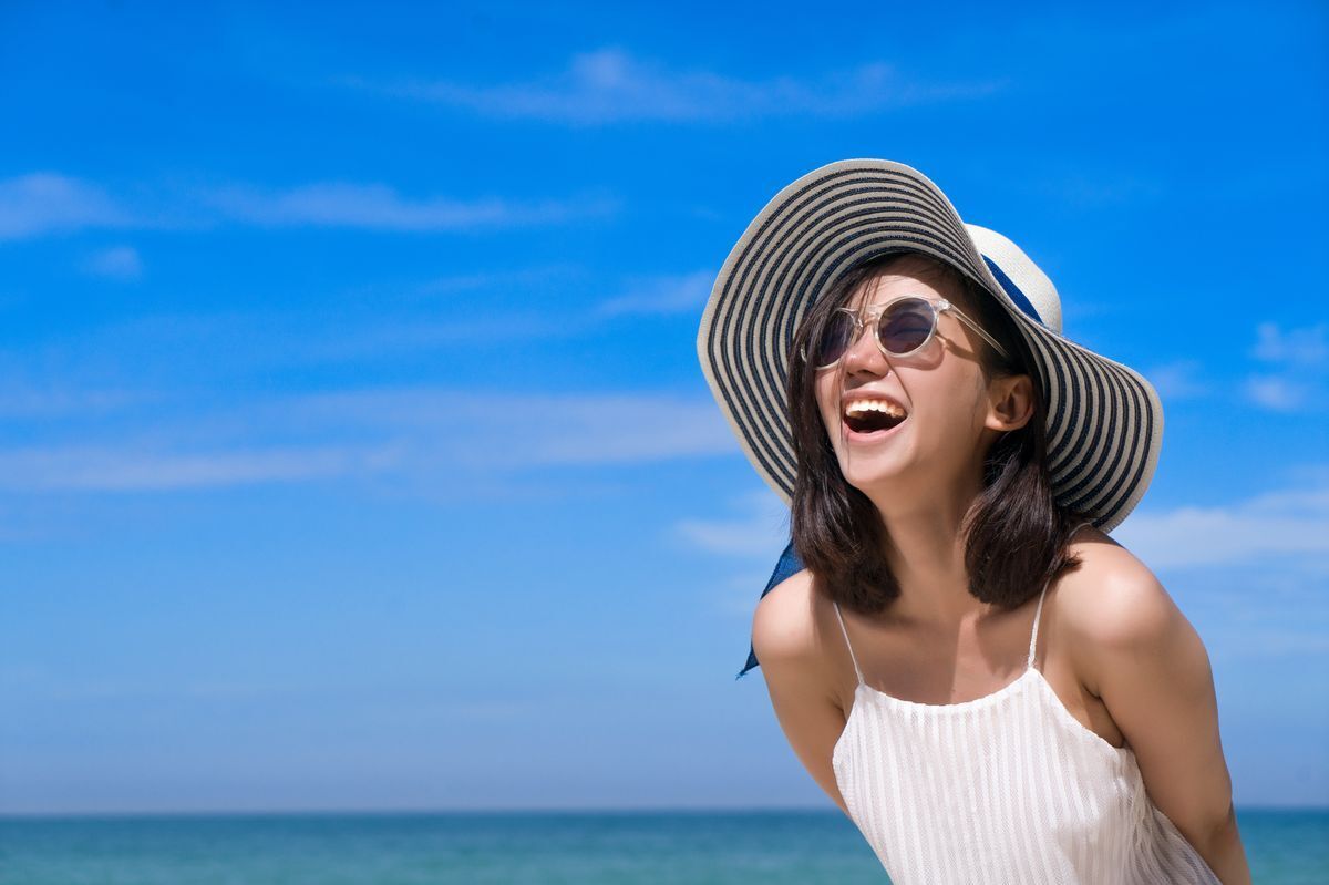Smiling Asian young woman at the beach with a sun hat and sunglasses.