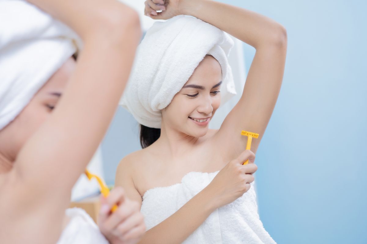 Woman wrapped in white towel shaving her underarm with a yellow razor. 
