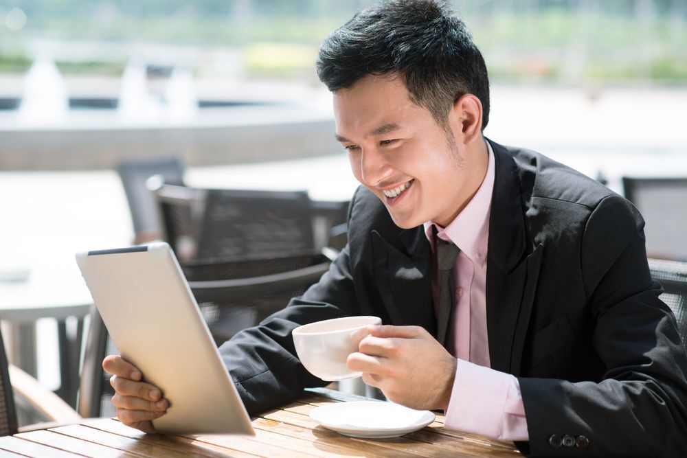 A young Asian man in a suit having coffee while looking at his tablet