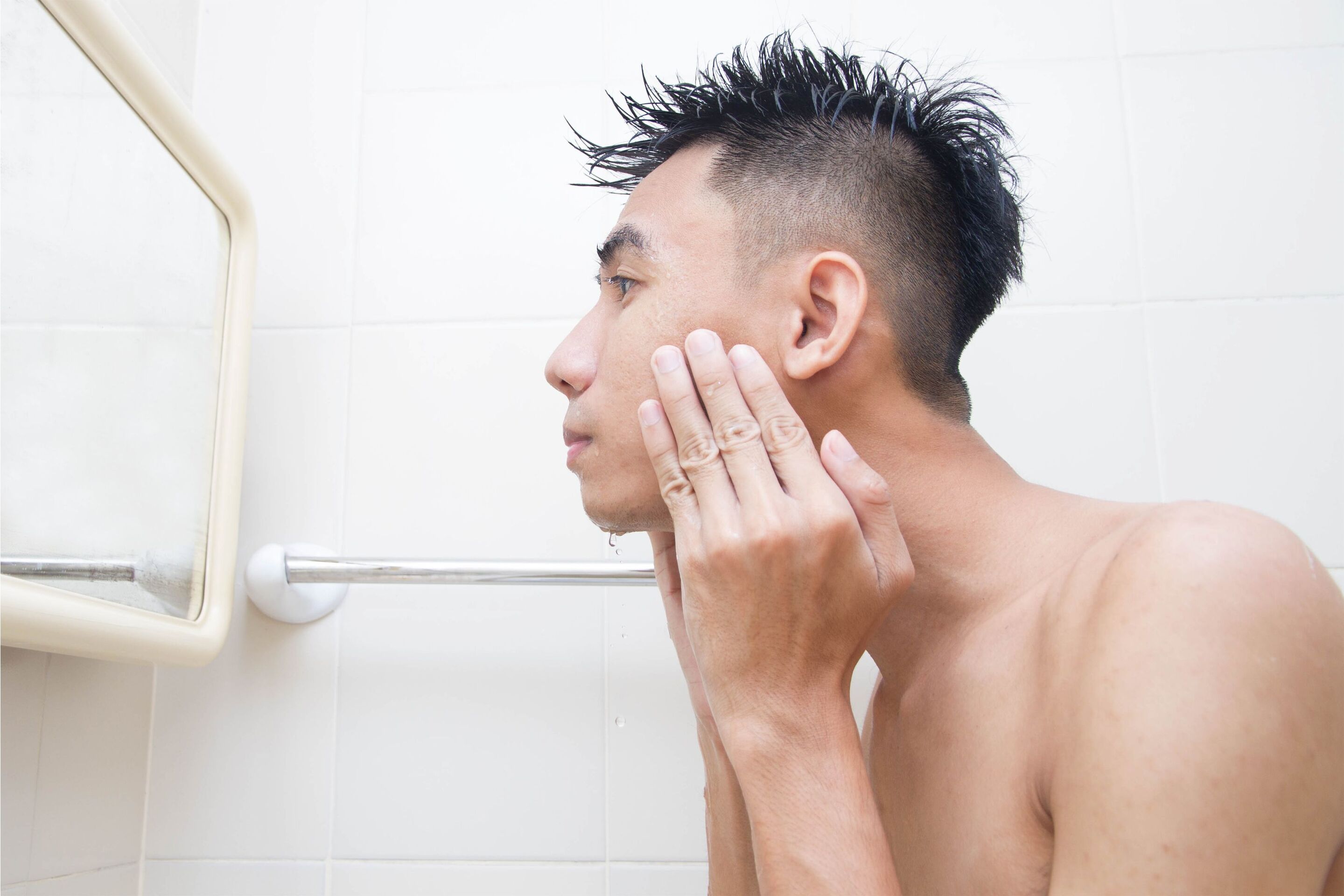 An Asian man washing his face in front of a mirror