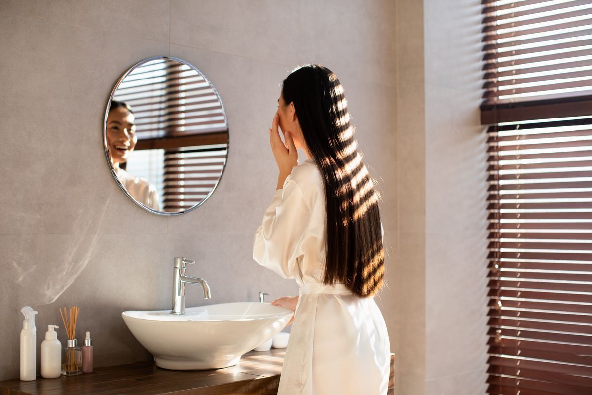 Asian woman with long hair in a bathrobe looking at herself in the mirror.