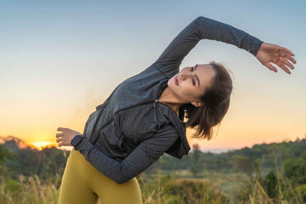 Woman in a gray jacket and yellow leggings stretching her body before a workout.
