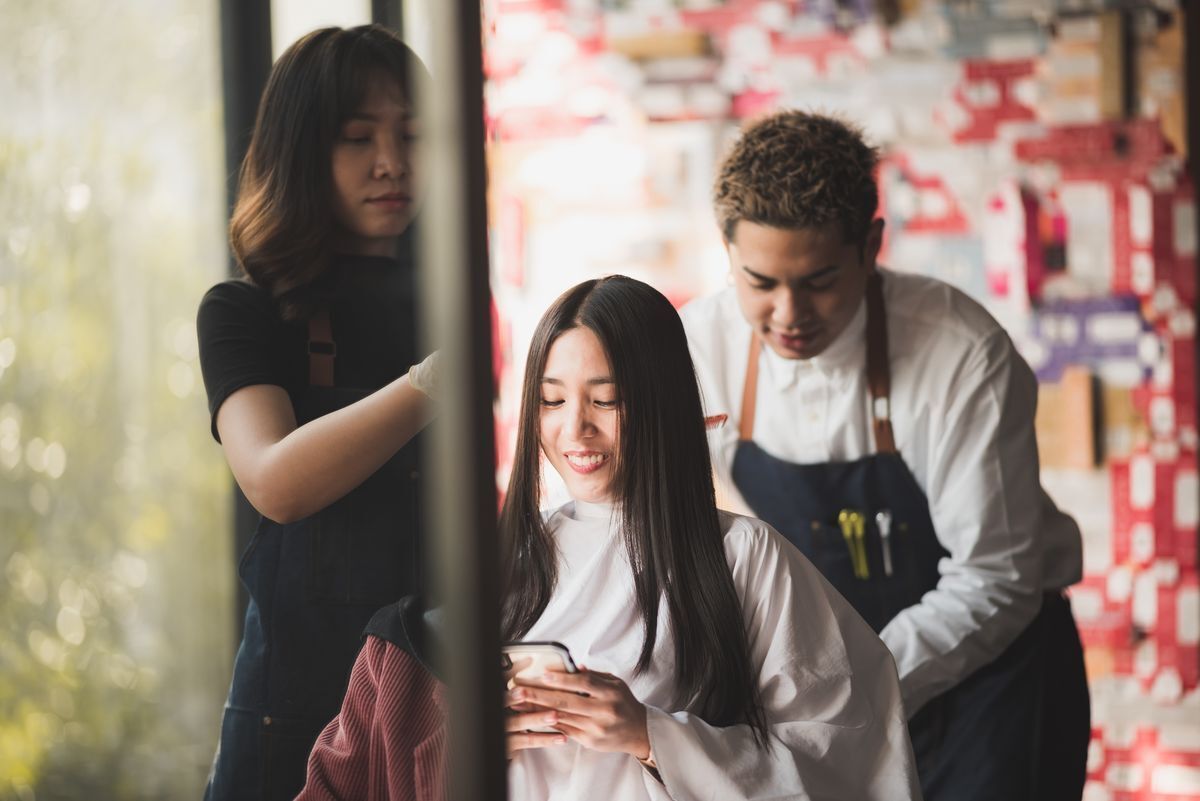 Smiling Asian female looking at her phone in the salon with female and male hair stylists.
