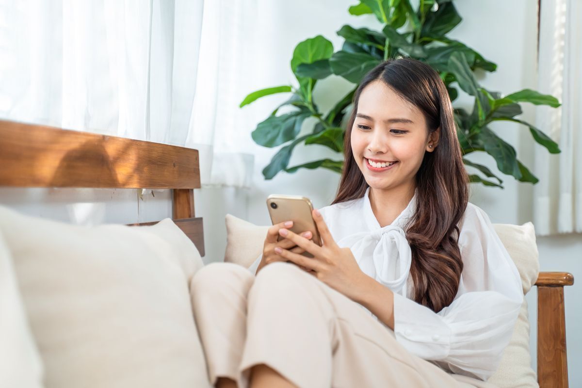 Smiling Asian woman seated on a sofa, browsing on her phone.