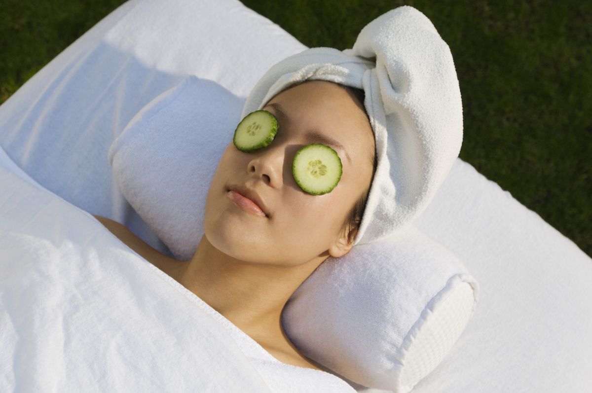 Woman lying down on a white spa bed with her hair wrapped in a towel and slices of cucumber on her eyes.