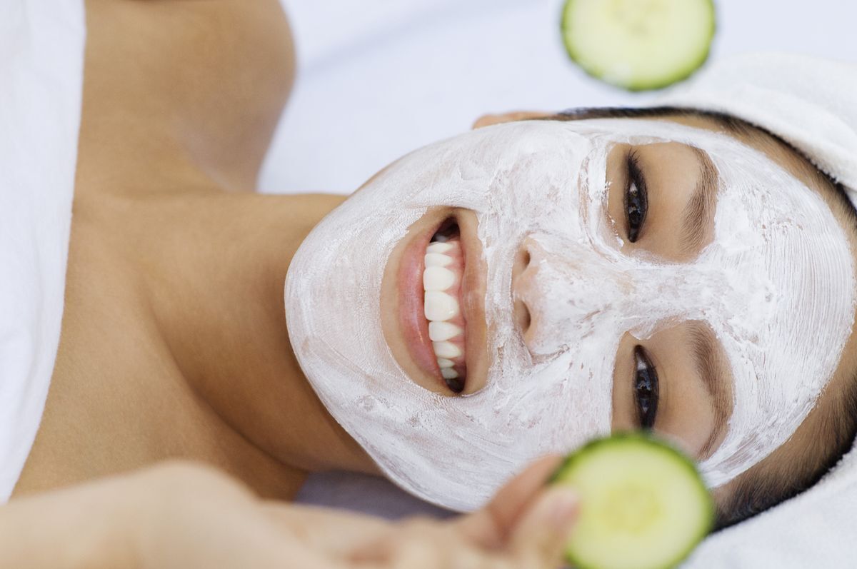 Close up of a smiling woman with a white face mask holding up a cucumber slice.  