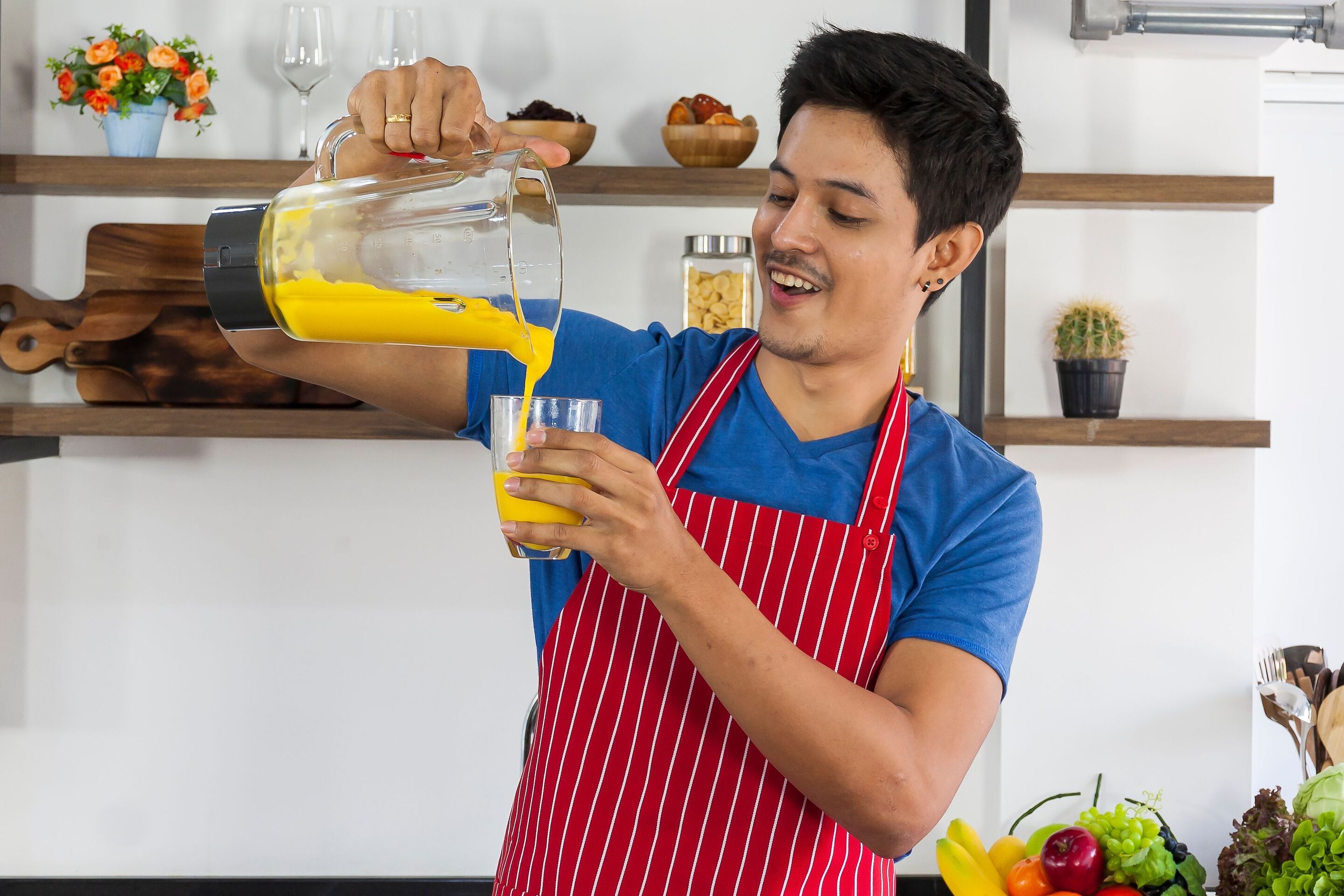 An Asian man wearing a red striped apron and pouring a yellow smoothie into a glass