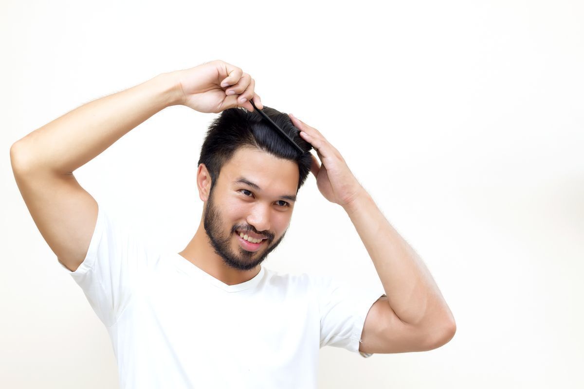 Smiling Asian man combing his hair wearing a white t shirt against a white background.
