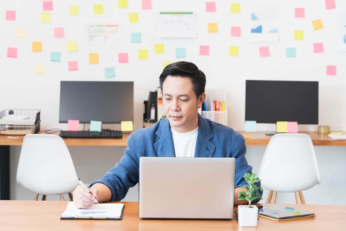 A middle aged man writing on his notebook while working in front of a laptop.