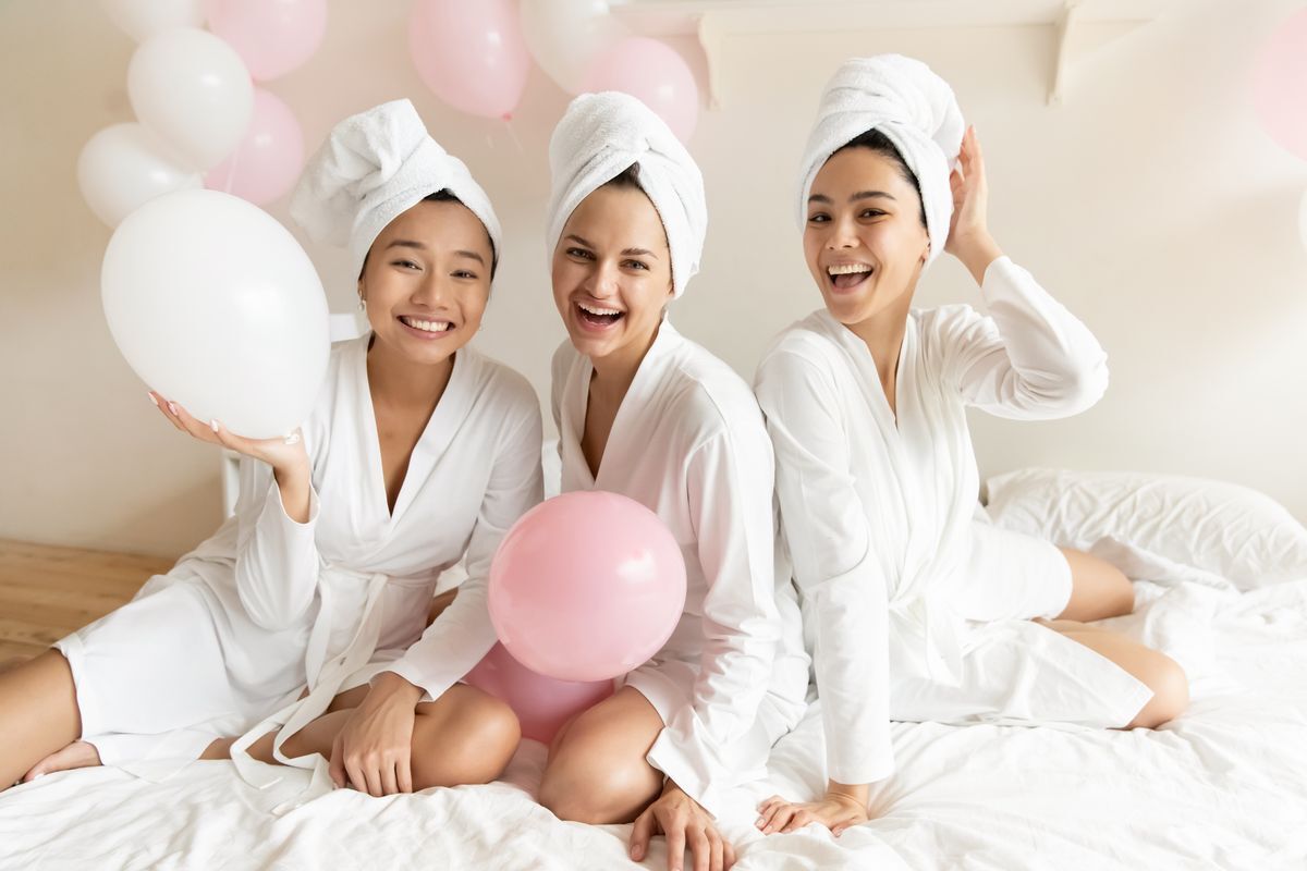 Three Asian women in bathrobes at a spa party, smiling while surrounded by pink and white balloons. 