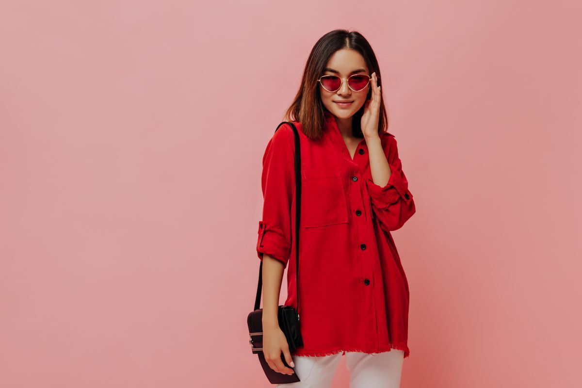 Young Asian woman with a handbag and sunglasses smiling against a pink background.