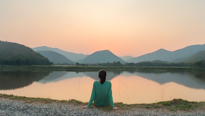Back of woman with dark hair staring into a lake with mountains surrounding her