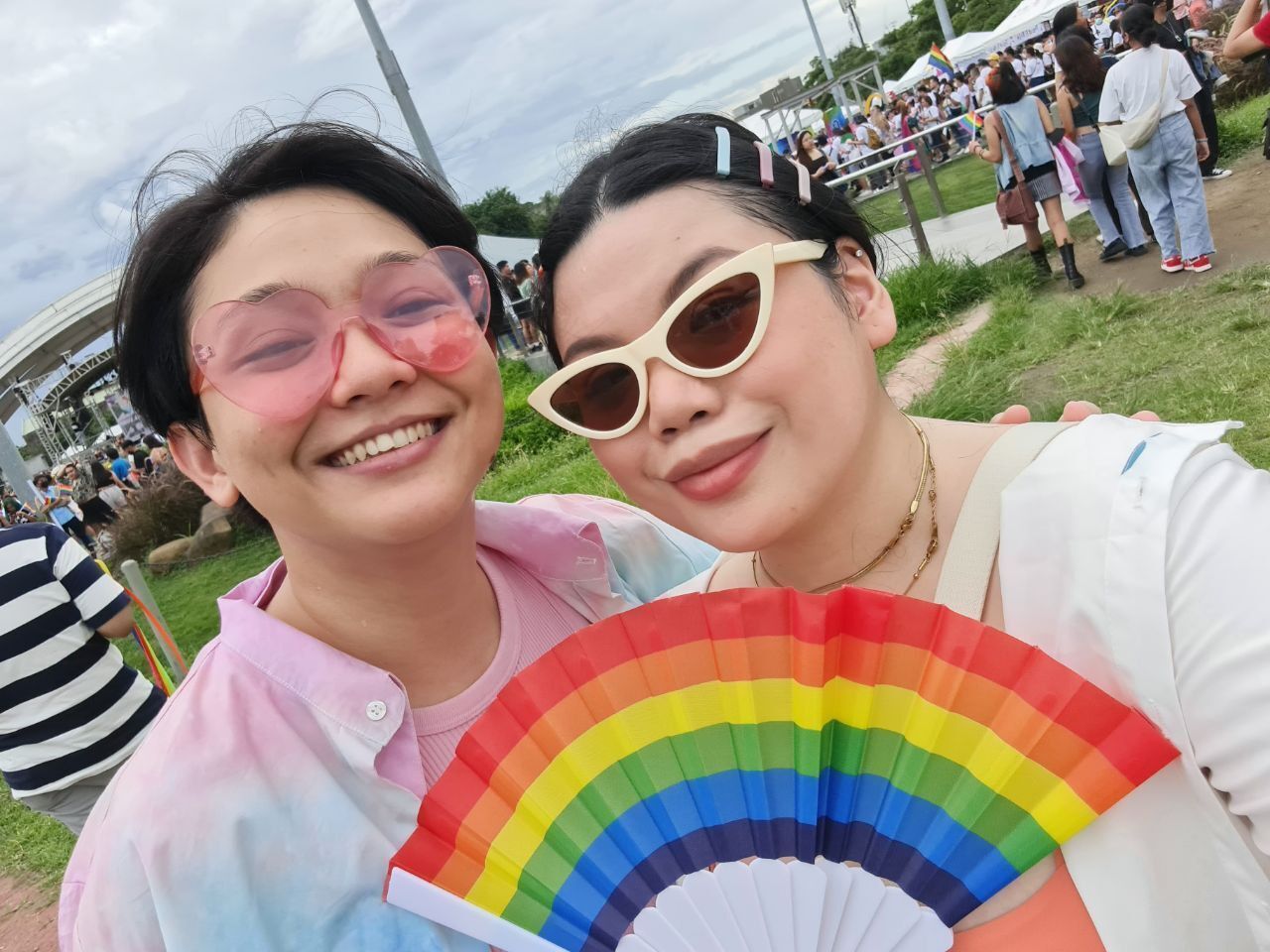Two women in an outdoor festival holding a rainbow colored fan.