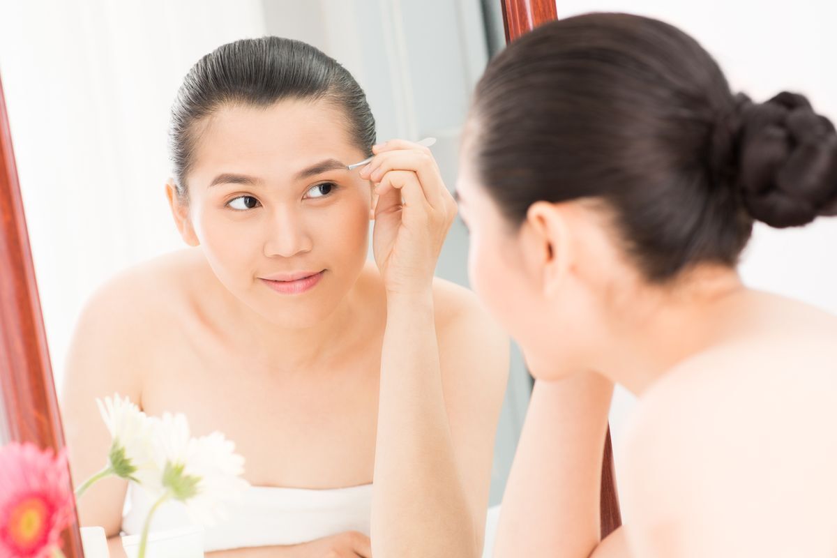Woman plucking her eyebrows with a pair of tweezers in front of the mirror.  