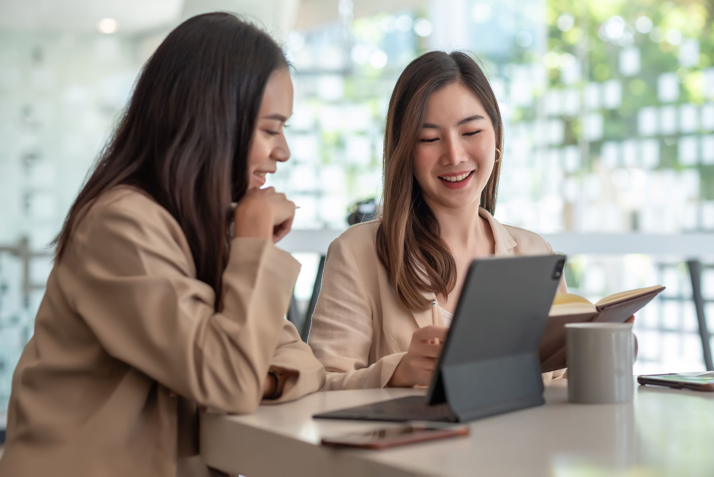 Two Asian women happily working together with a tablet and notebook.