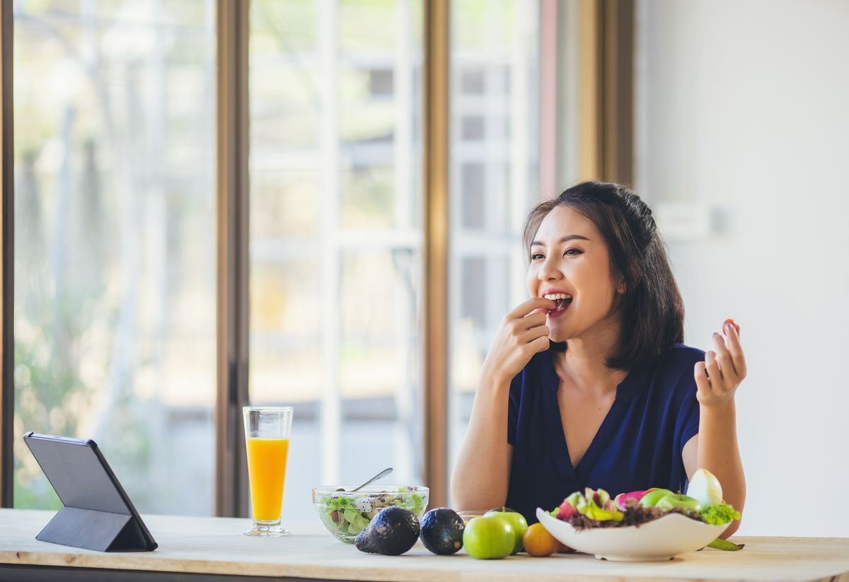 Asian woman snacking on fruits and vegetables in front of her tablet