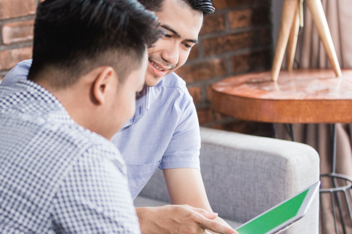 Asian men sitting on a couch with one pointing at a tablet and smiling.