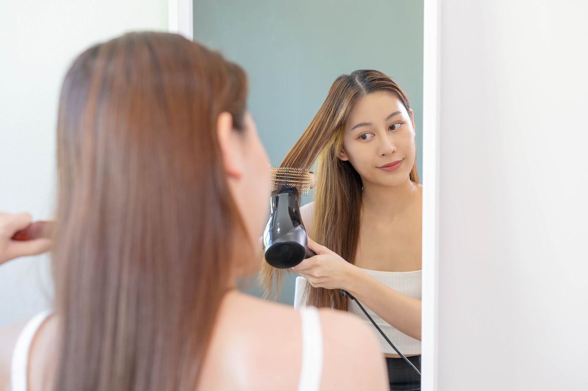 Asian woman blow drying her hair in front of a mirror