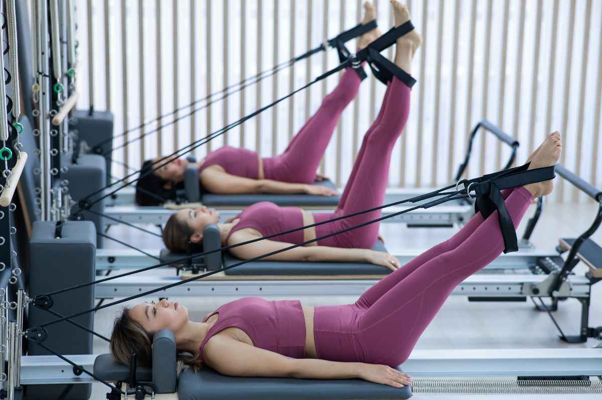 Three women in purple workout clothes stretch their legs on a reformer.