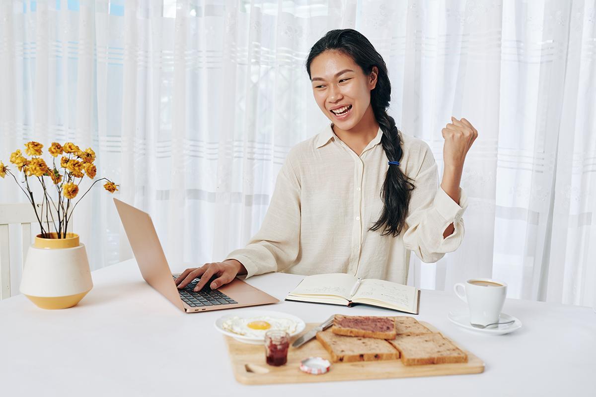 Asian woman pumping fist in the air while looking at laptop and eating breakfast