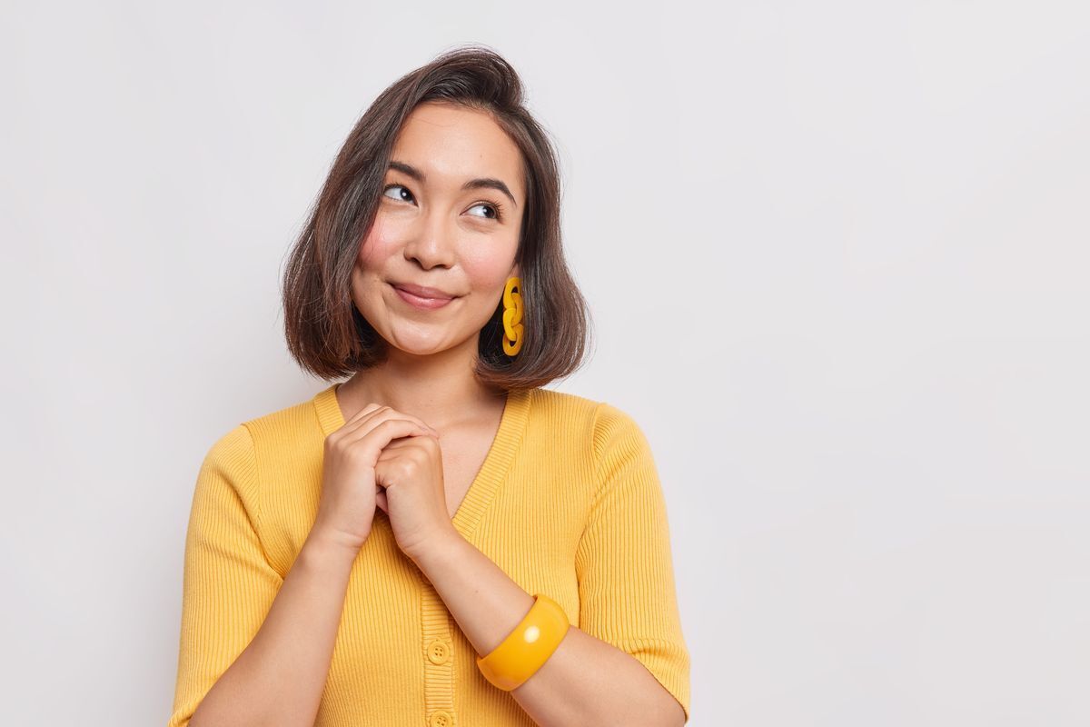 Asian woman wearing a yellow shirt, yellow earrings, and a yellow bangle bracelet smiling and looking up against a white background.