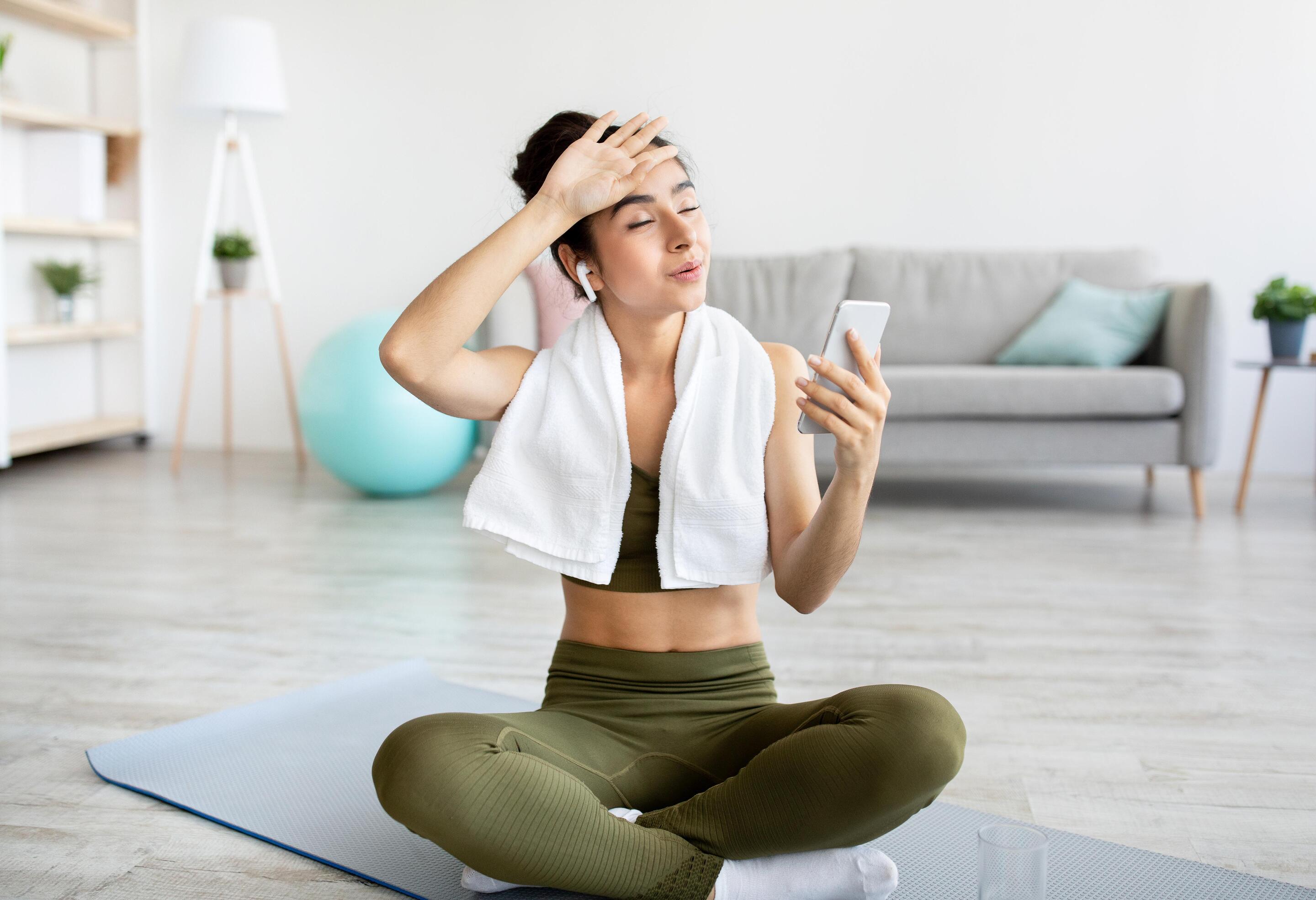 Young Asian woman in olive green workout clothes with hand on forehead and towel and cellphone