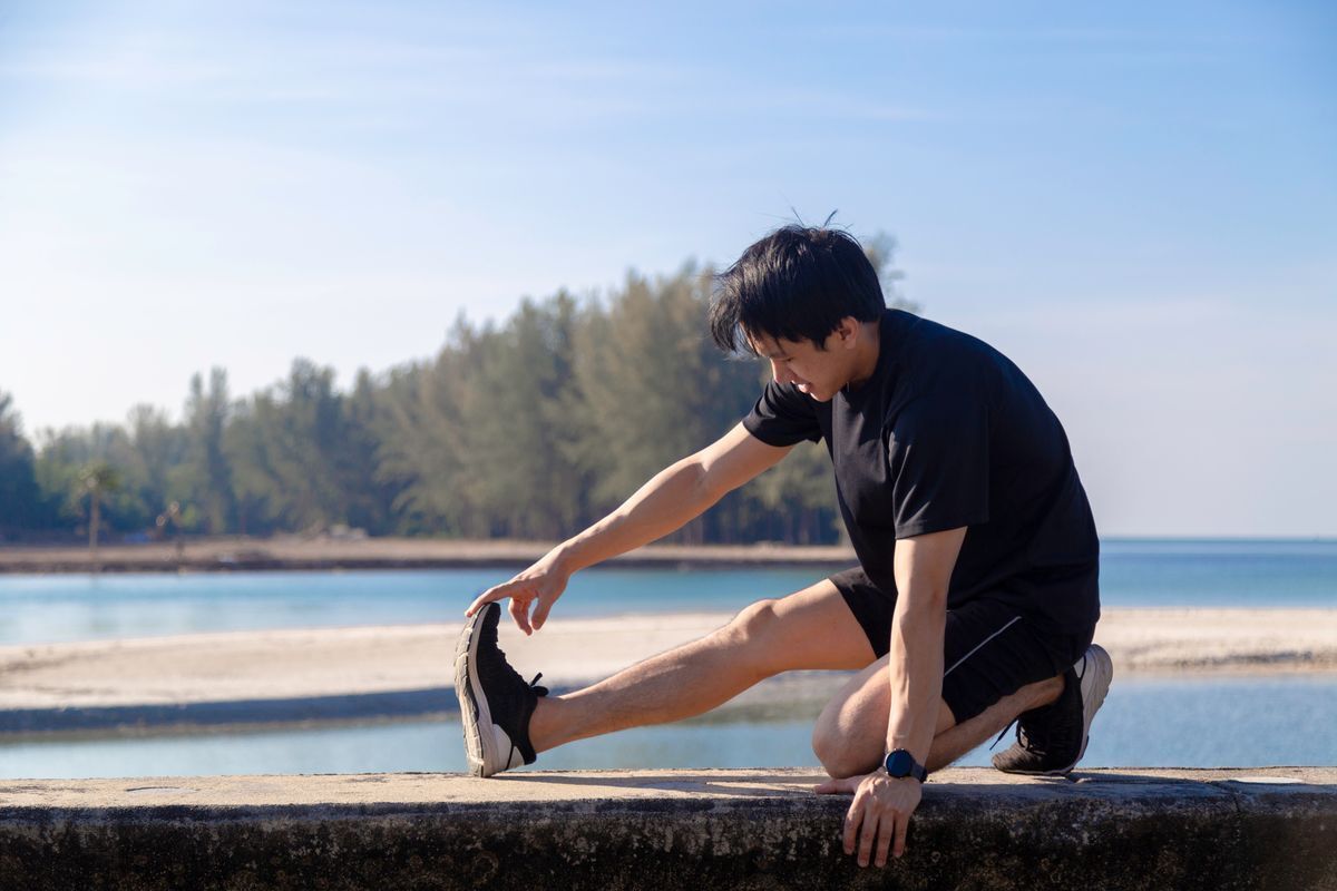 Asian man doing a leg stretch exercise outdoors with a view of a lake.
