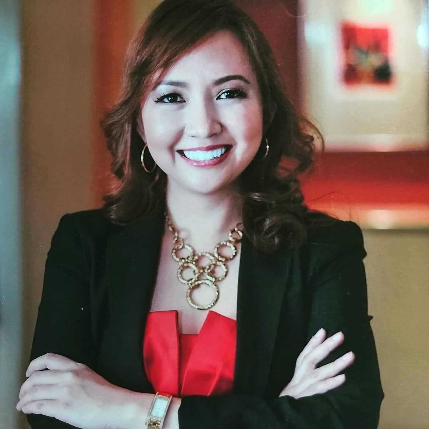 Portrait of a woman in a blazer and chunky jewelry smiling with her hands folded. 