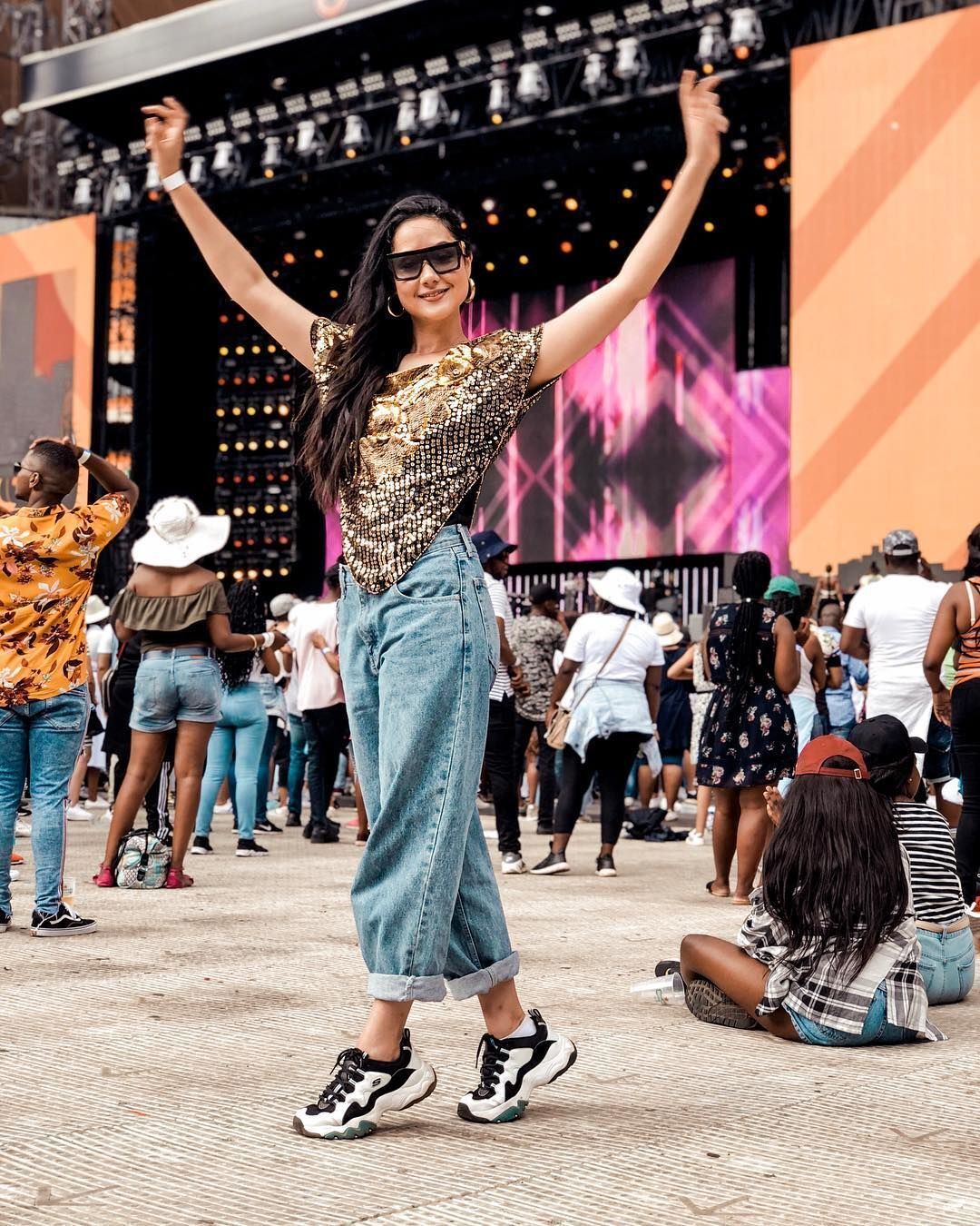 A portrait of woman wearing a sparkly top, jeans, sneakers, and glasses at a music concert.