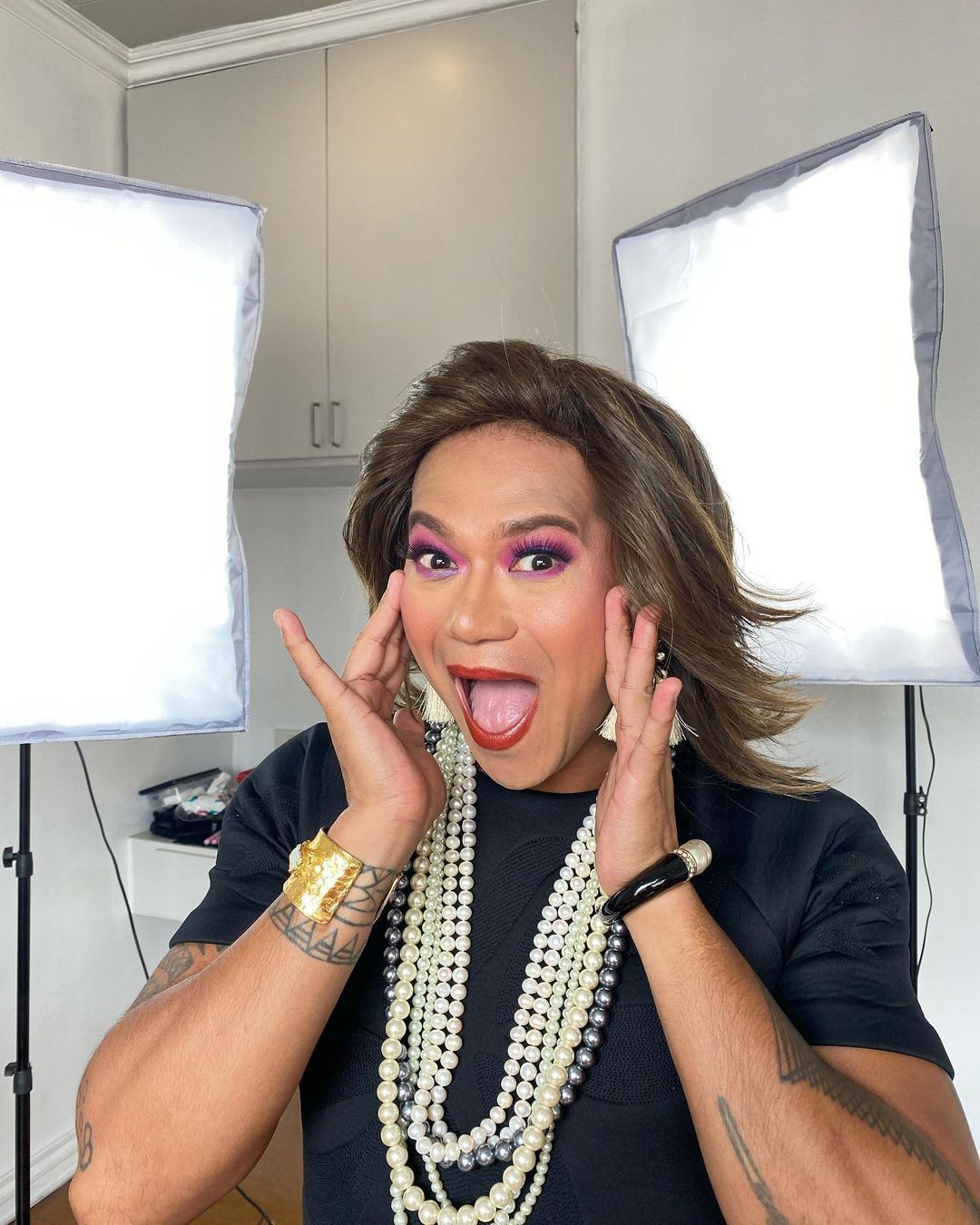 Lourd Ramos wearing a brown wig, black shirt, and layers of pearls