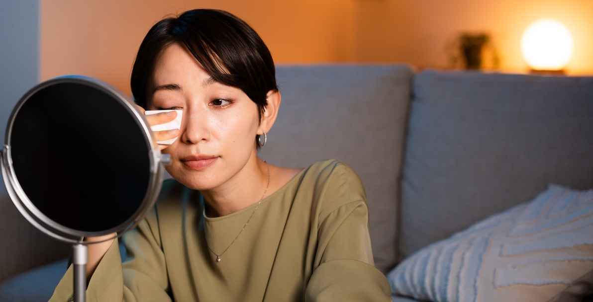 An Asian woman removing her eye makeup with a cotton pad in front of the mirror.