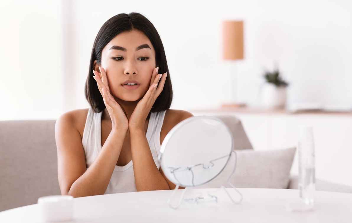 An Asian woman applying skincare in front of a mirror.