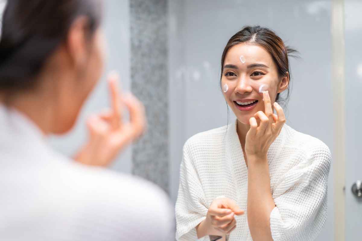 A smiling Asian woman applying moisturizer to her face in front of a mirror.