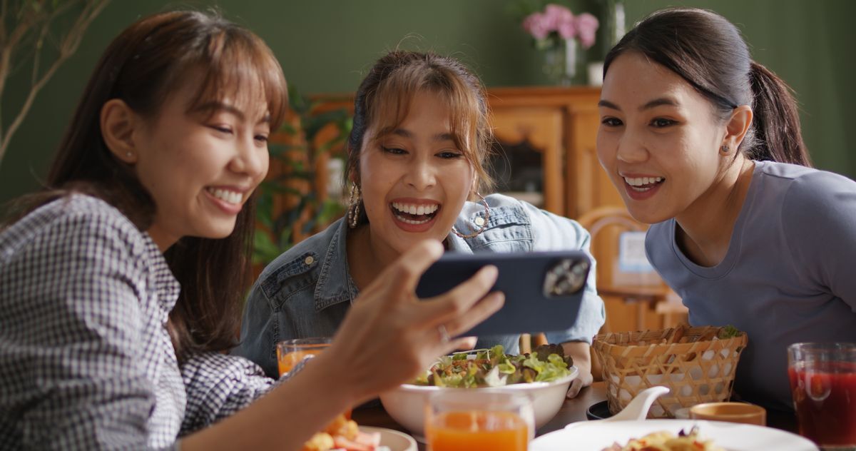 Three Asian woman hanging out over a meal while watching something on a phone.
