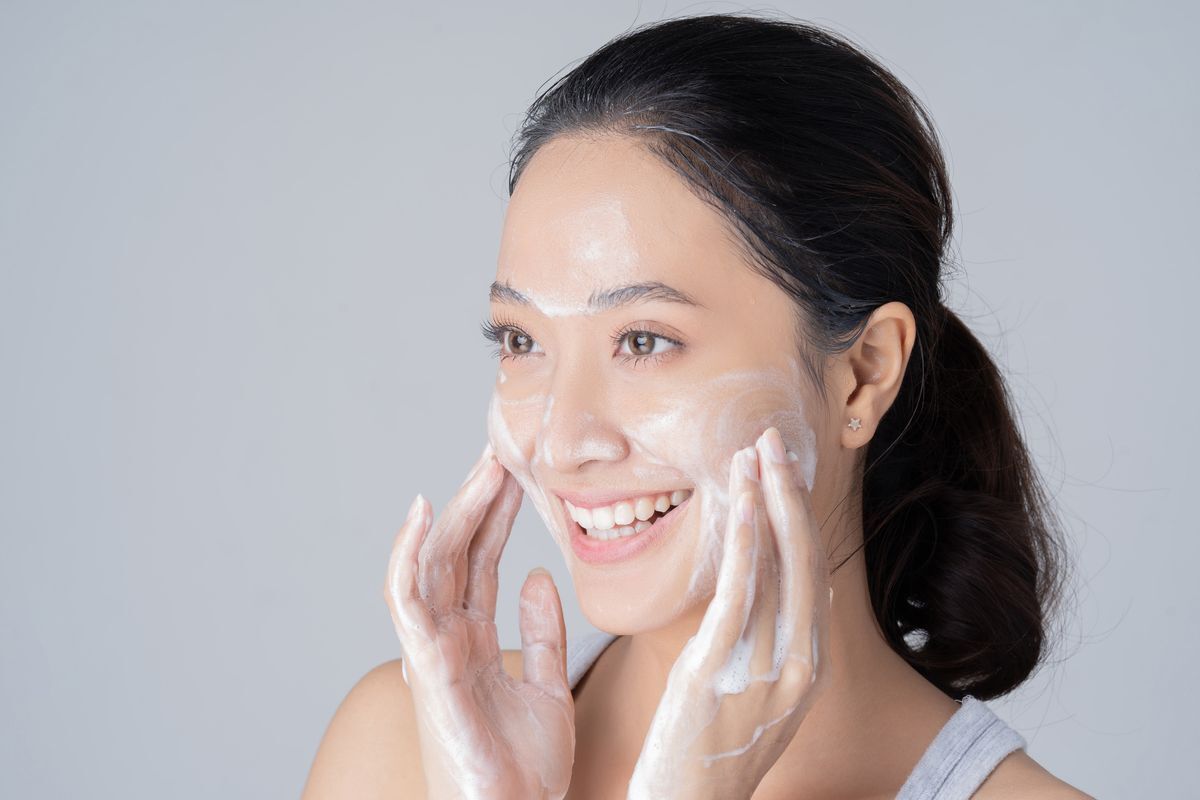 Young Asian female happily washing her face with soap against a gray background. 