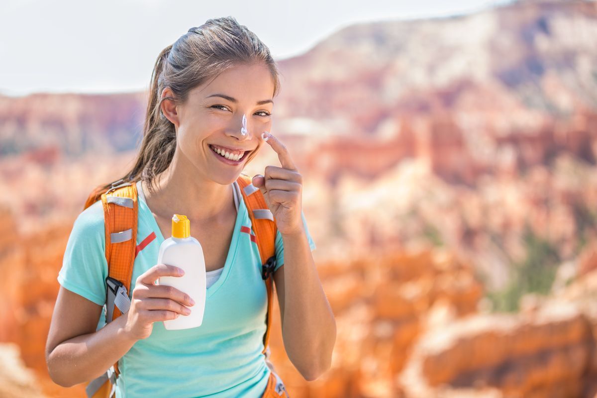 Portrait of a young female happily putting sunscreen on her nose against a blurred outdoor background.