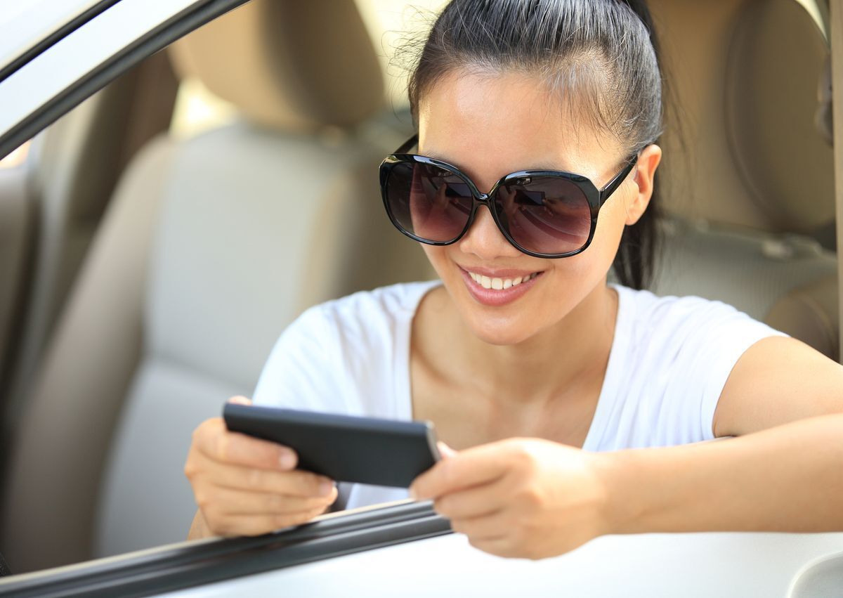 Asian woman wearing a ponytail and sunglasses sitting in a car while looking at phone