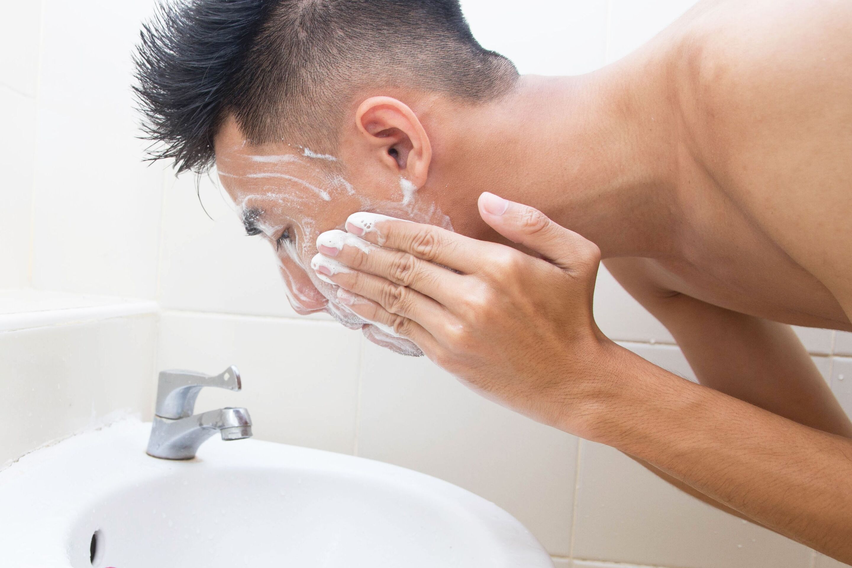 An Asian man washing his face with facial wash over the sink