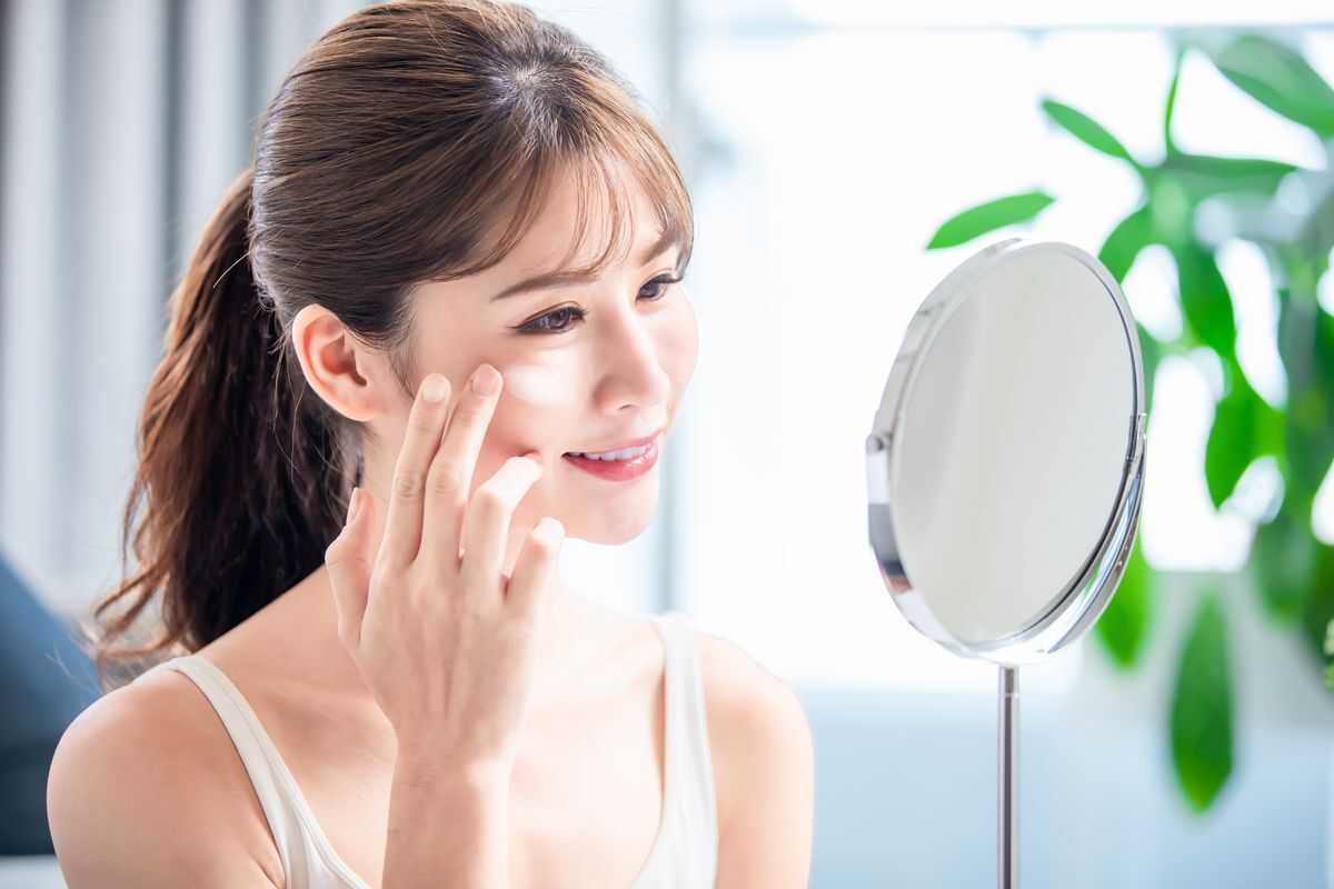 An Asian woman applying moisturizer in front of a desk mirror. 