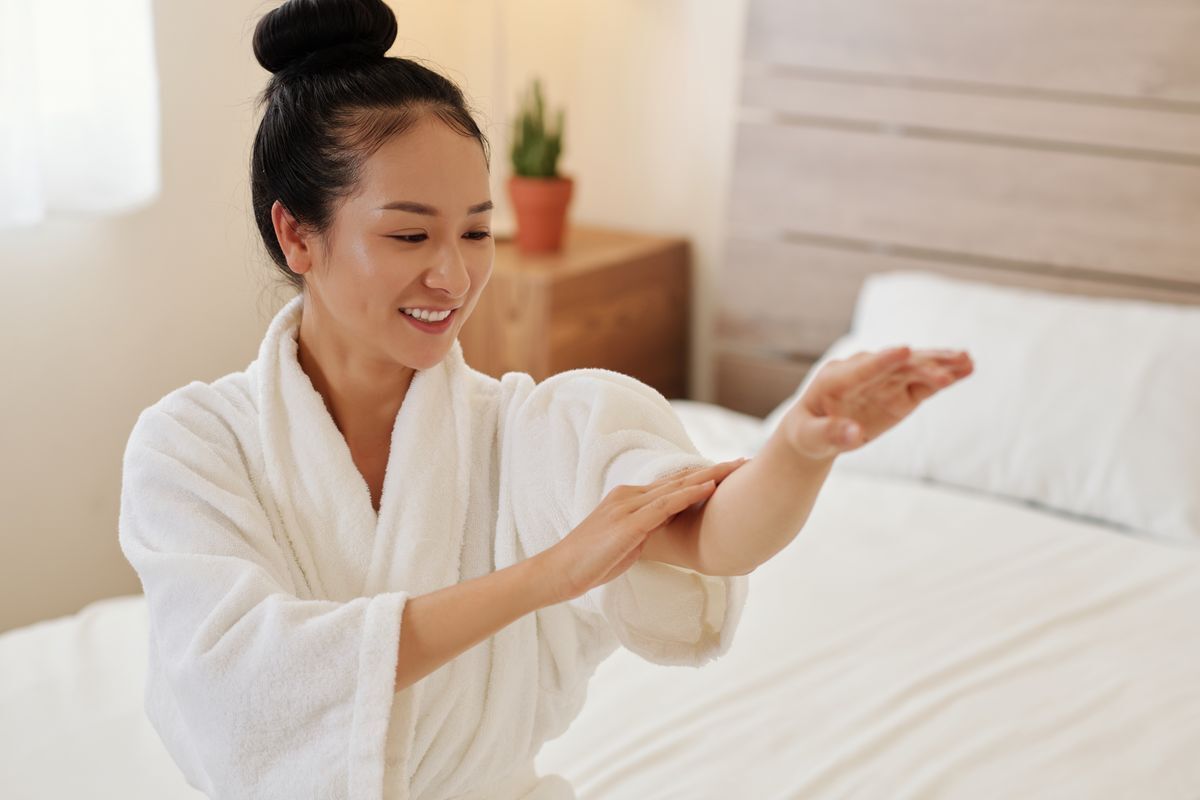 Smiling Asian woman wearing a white robe and touching her arm in her bedroom.