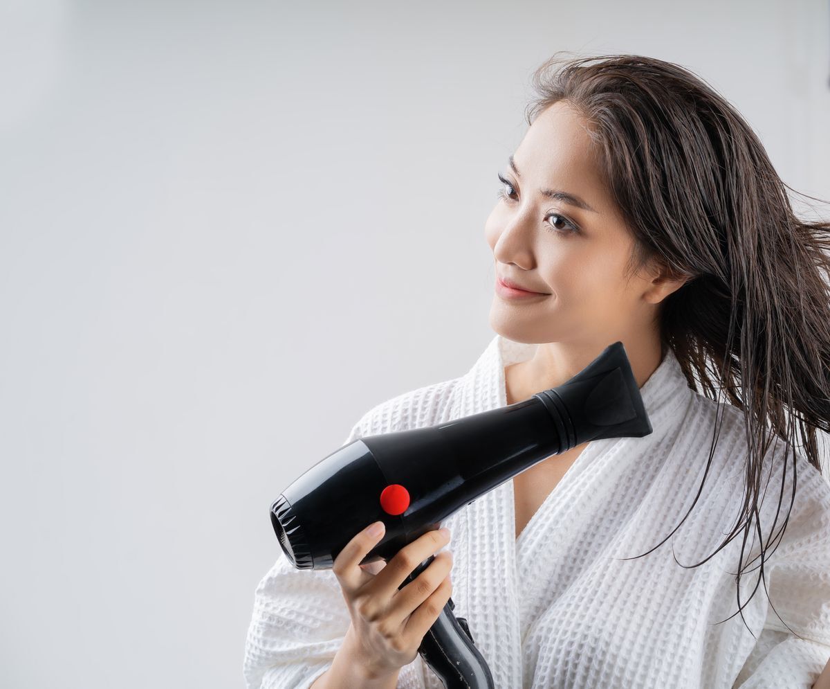 Smiling Asian woman in a white robe drying her hair against a white background. 
