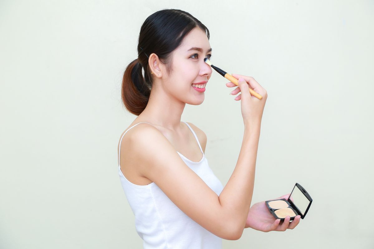 Side view of Asian woman smiling and applying makeup on her nose with a makeup brush against a white background.