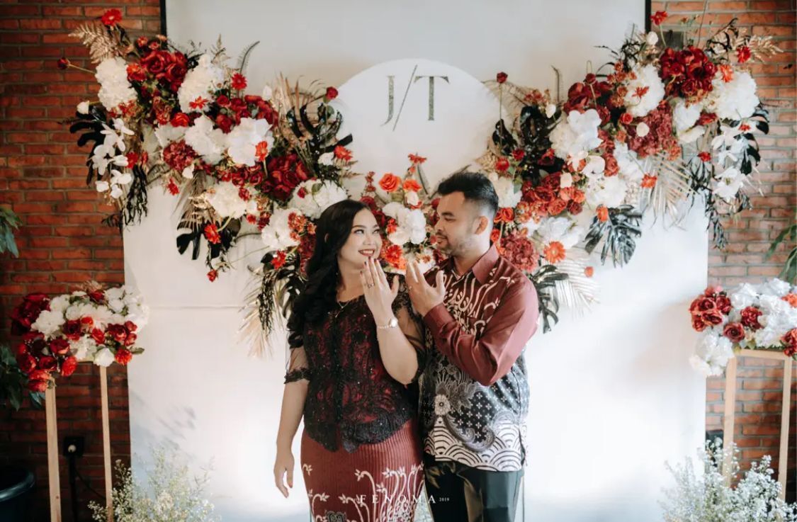 A portrait of smiling couple posing in front of a red and white floral backdrop.