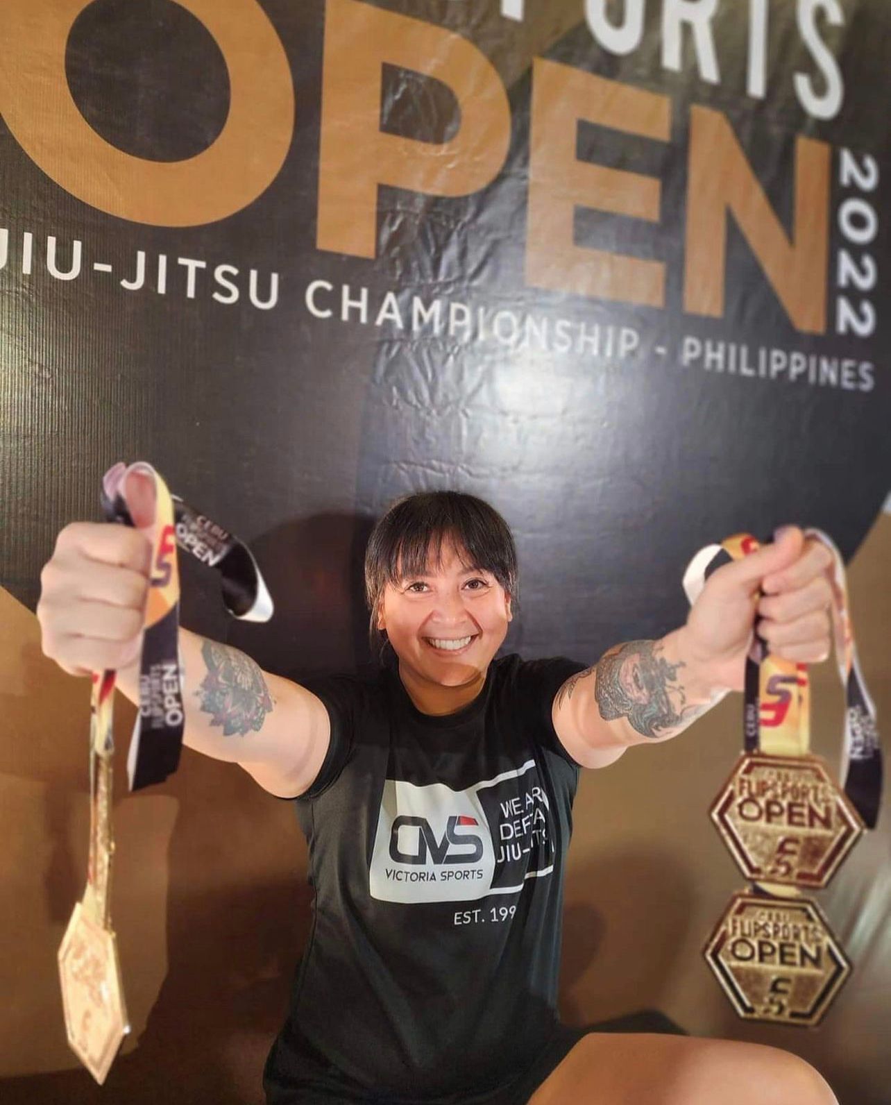 Asian woman in black smiling and holding medals against a black and brown background.