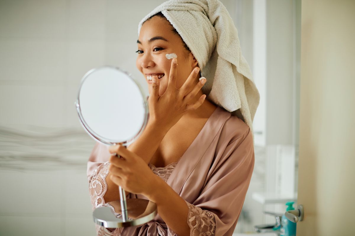 Asian woman applying cream on her face while holding a mirror.