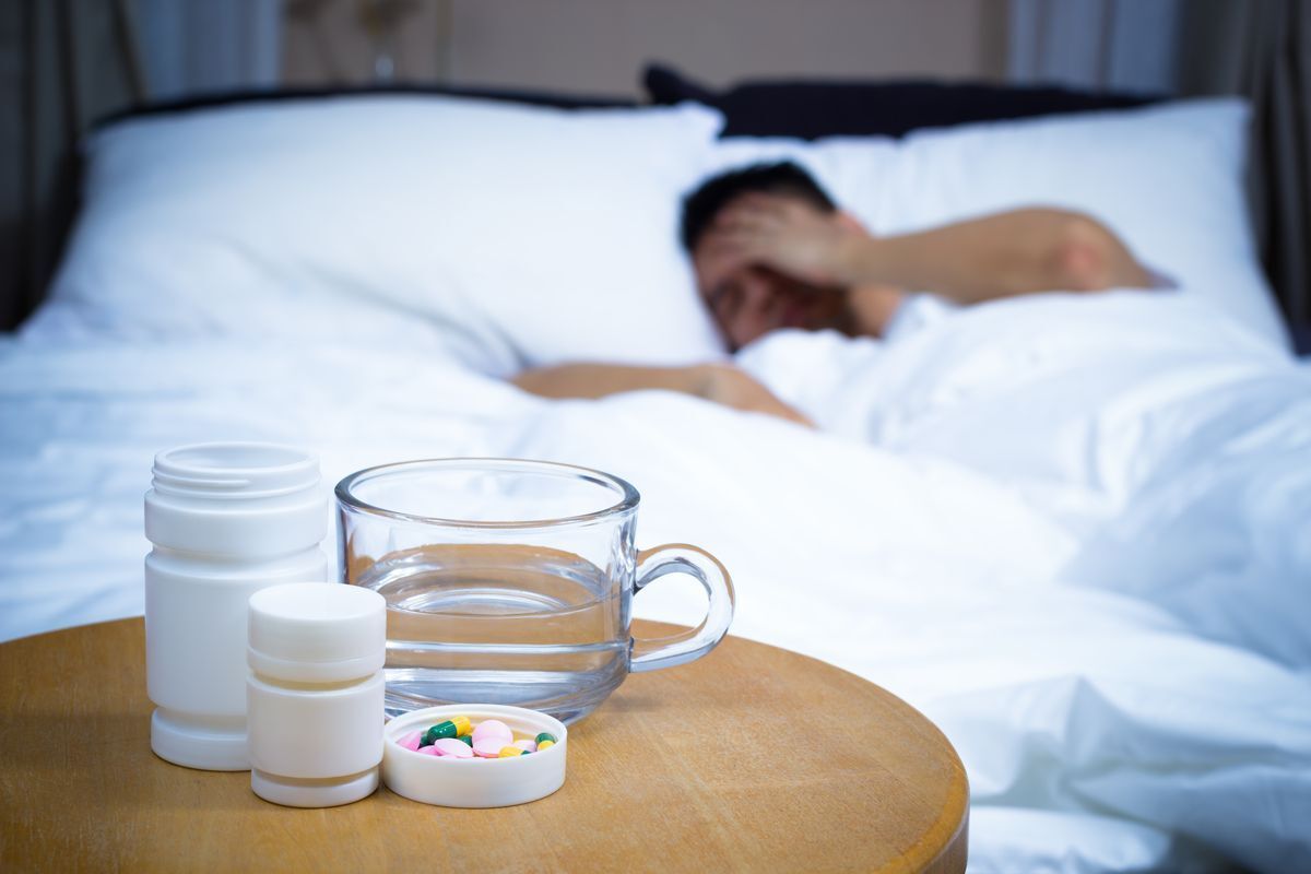 A man with a pillow over his head reaches for his medicine