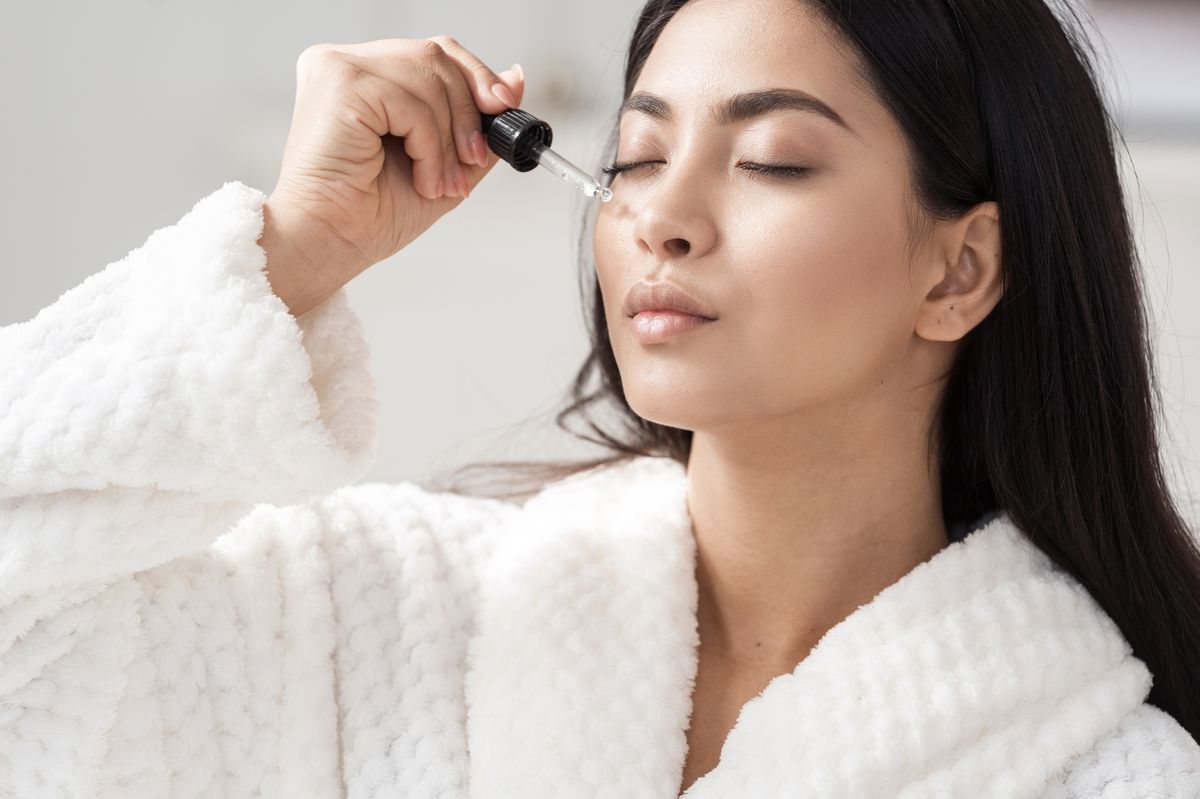 Woman in a white bathrobe applying serum on her skin.