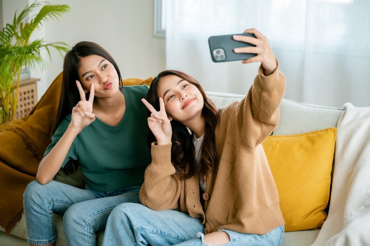 Two Asian women flashing peace signs and posing for a selfie on the sofa.