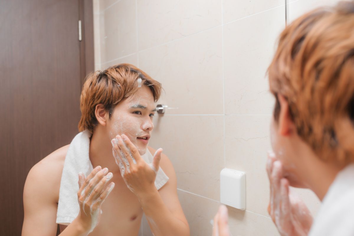 Asian man in the bathroom; looking at the mirror, exfoliating his face.