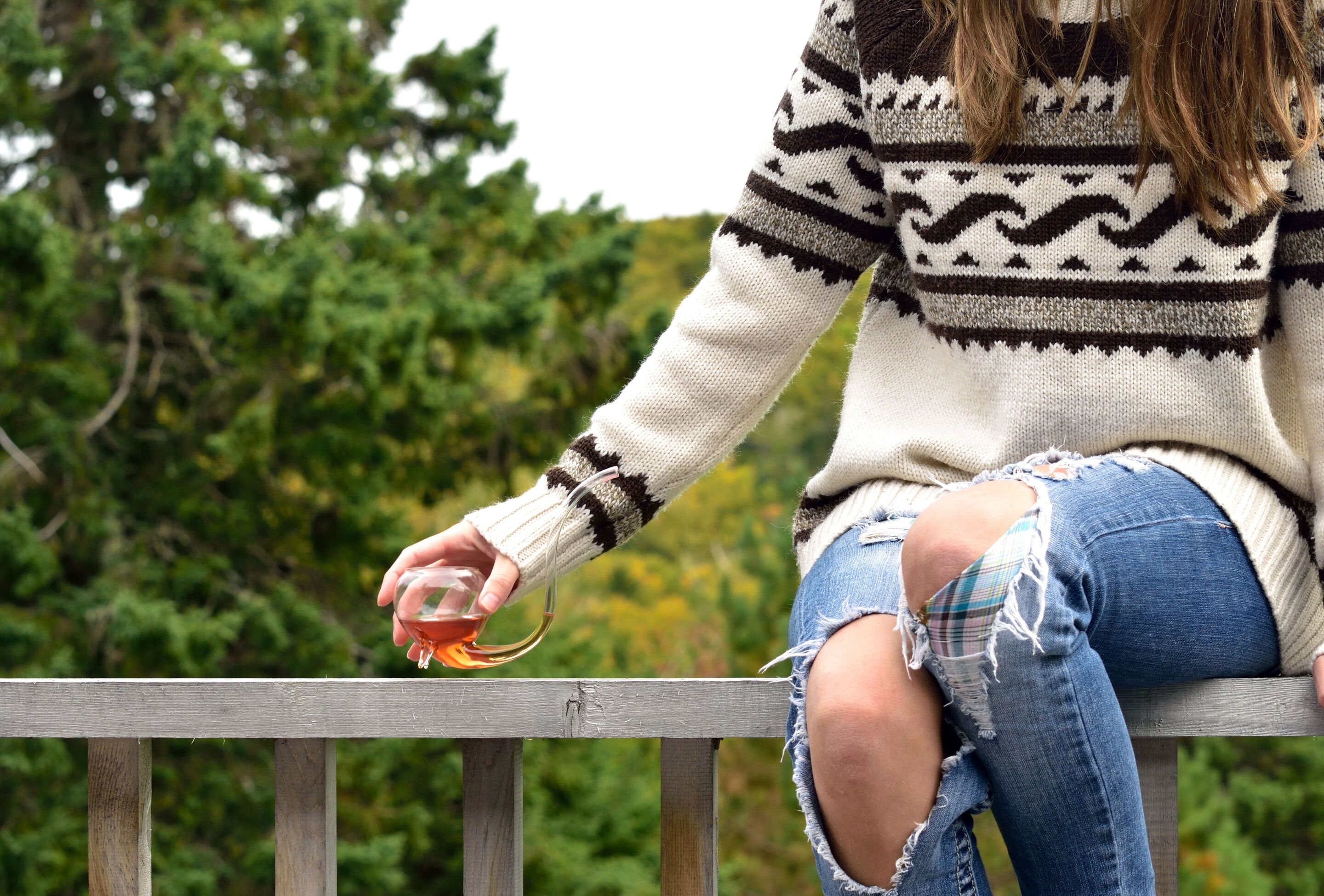 Asian woman wearing distressed jeans is sitting on a railing with a drink in hand